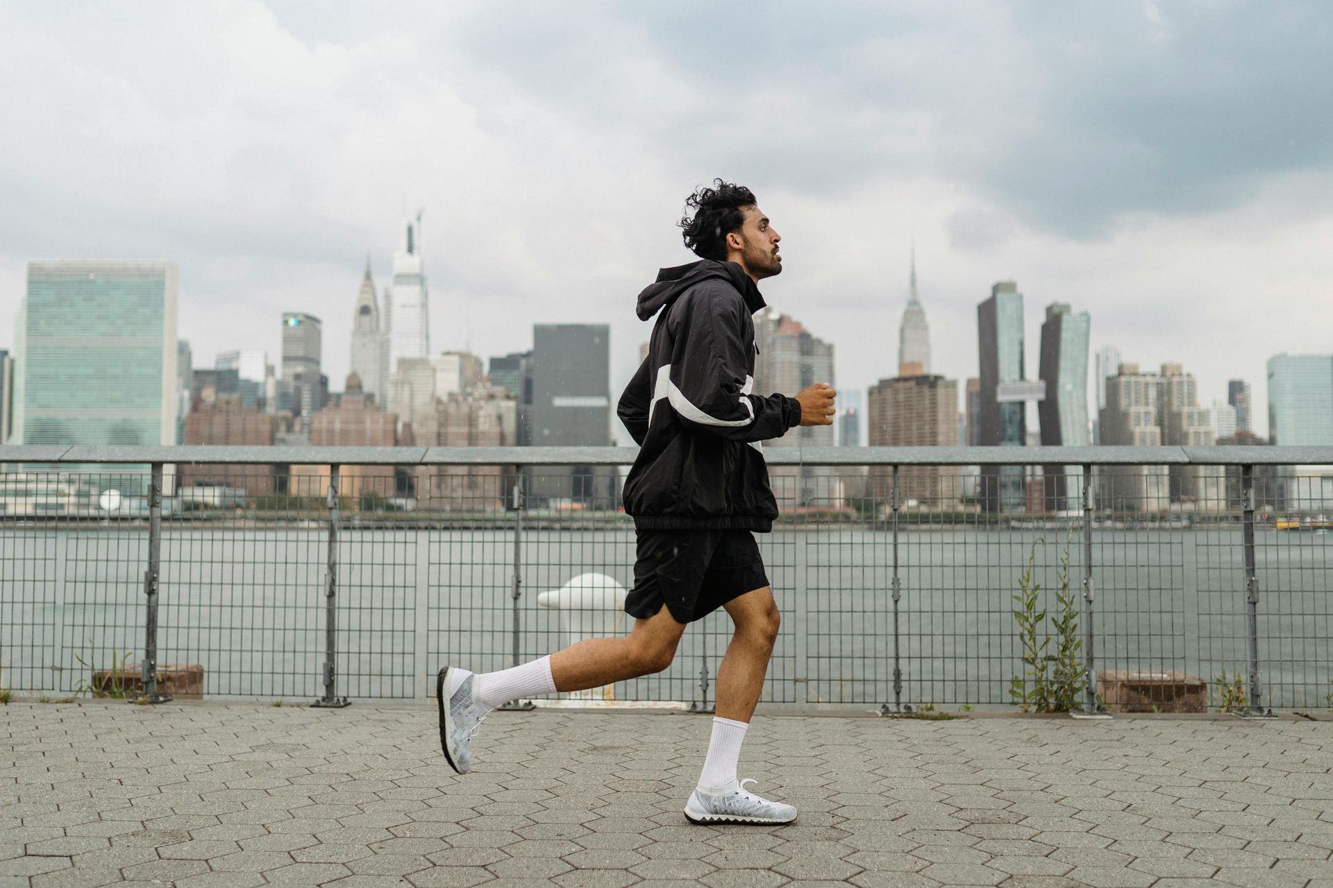Man running on a waterfront path with city skyline background under a cloudy sky.