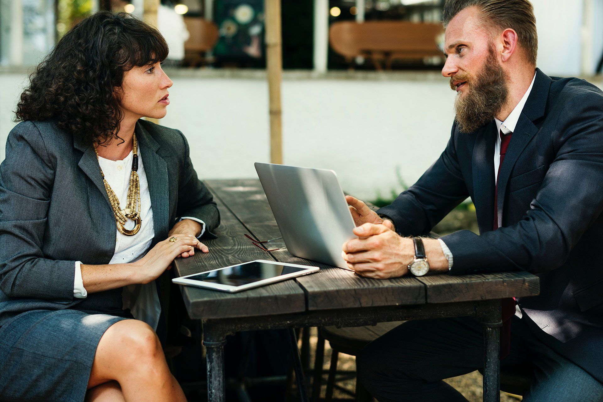 Woman and man in suits, looking at laptop and tablet on wooden table, discussing outdoors. Woman and man in suits, looking at laptop and tablet on wooden table, discussing outdoors.