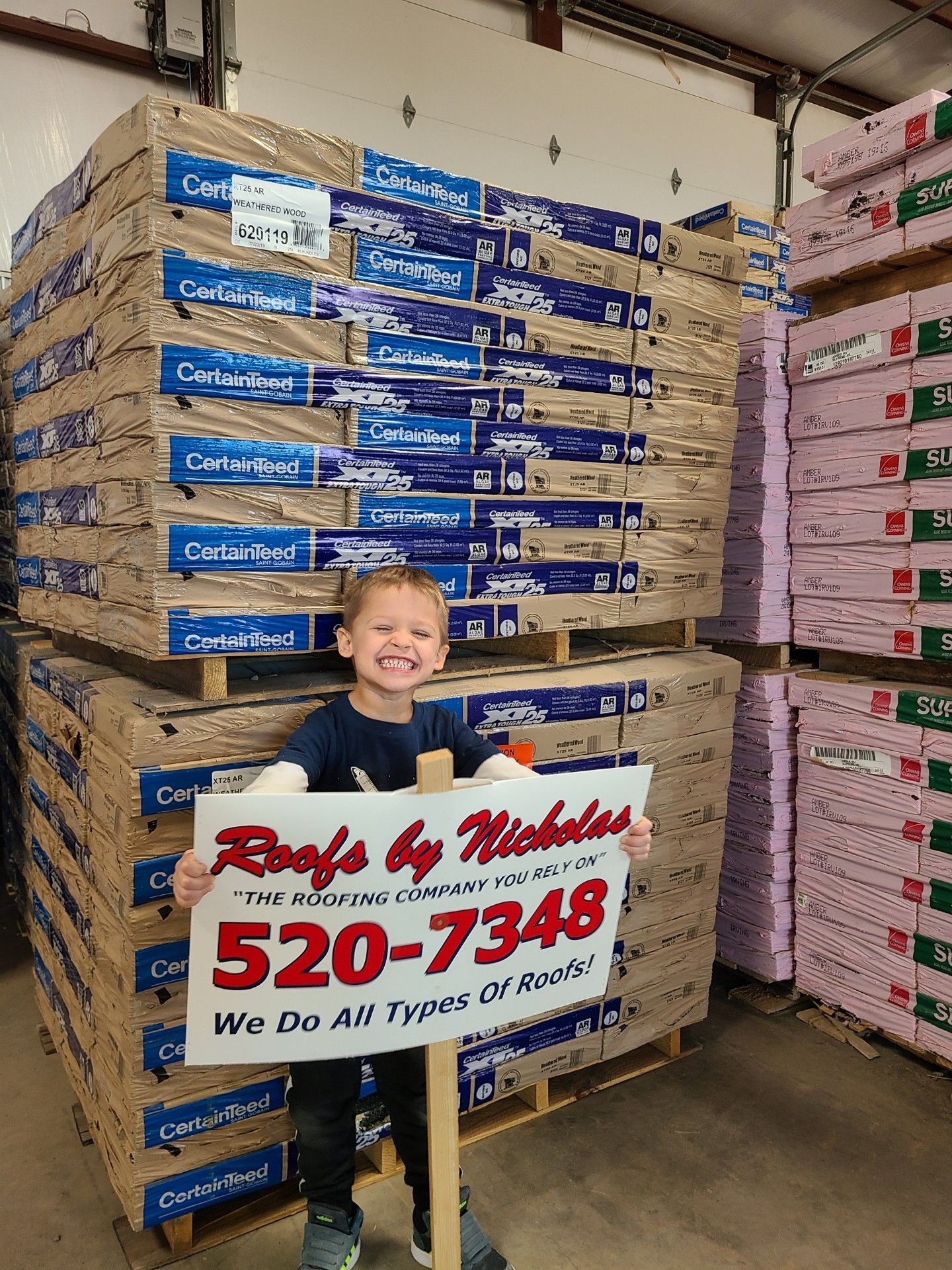 Smiling child holding Roofs by Nicholas sign in a warehouse with stacks of CertainTeed roofing materials