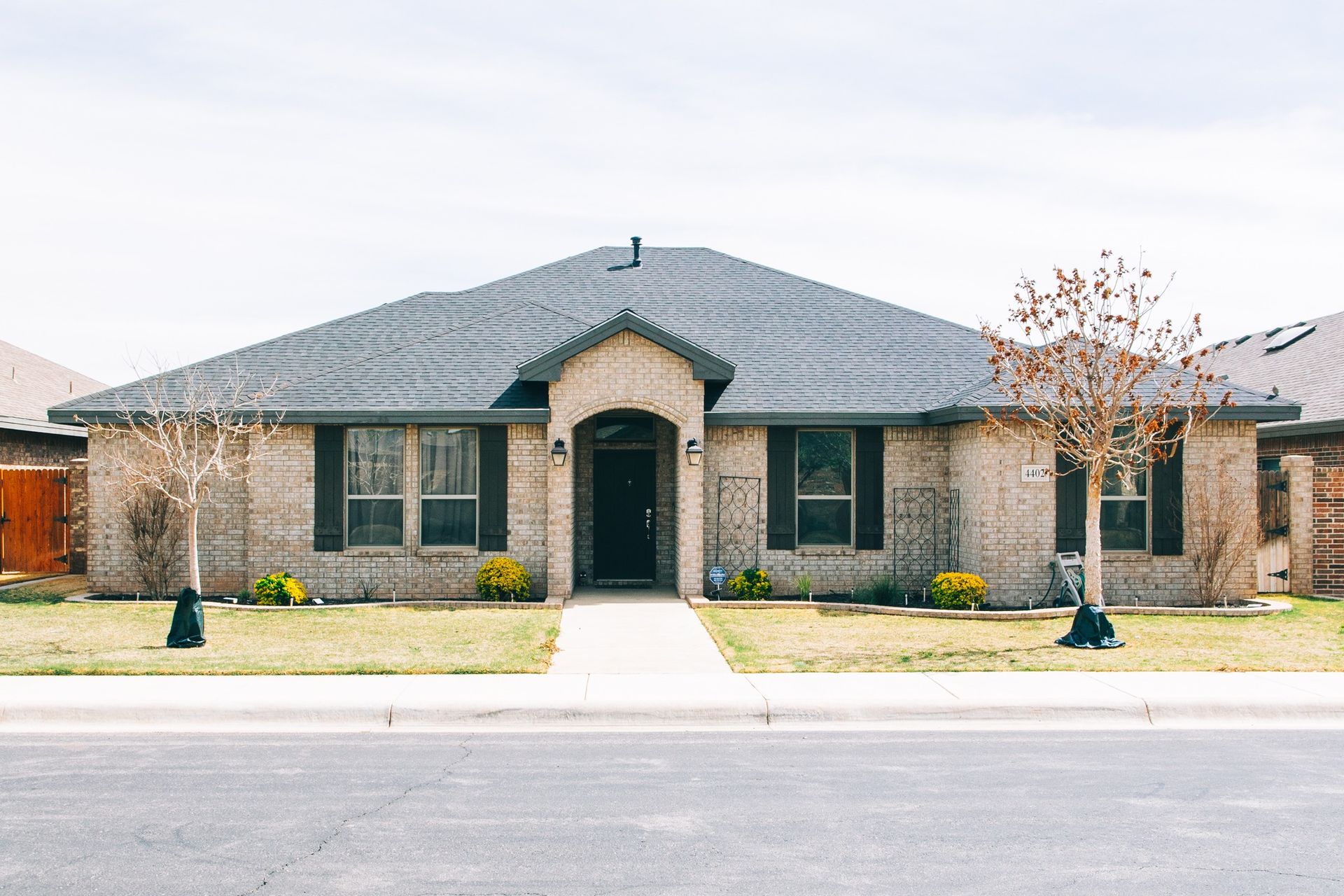 Single-story light brick house with dark gray shingle roof and arched entryway, photographed from street