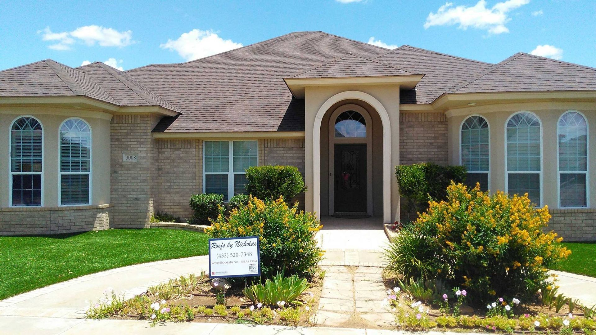 Roofs by Nicholas sign in front yard of beige brick house with brown shingle roof under blue sky