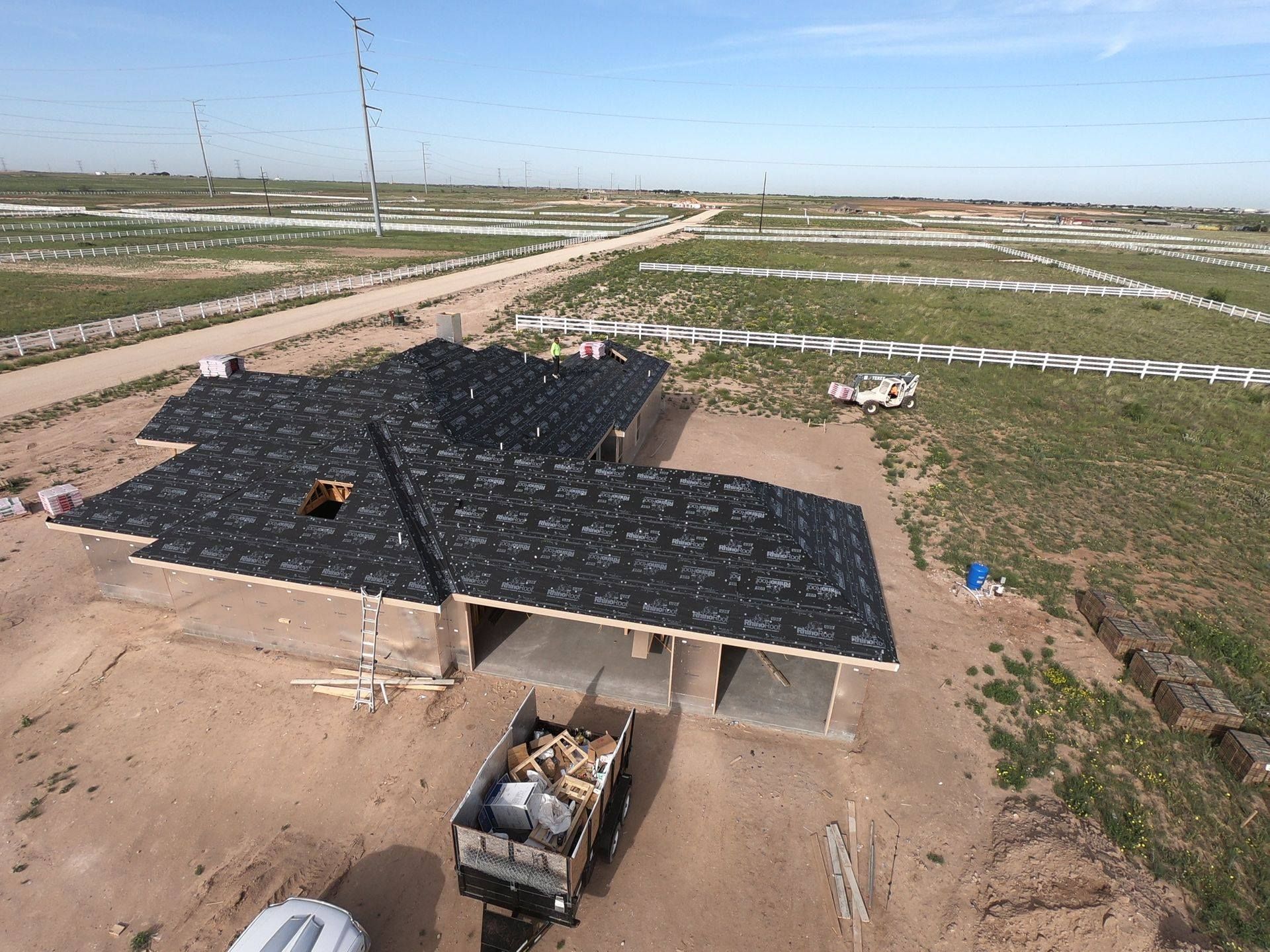 New house under construction with black roofing underlayment installed, surrounded by open grassy fields and dirt roads