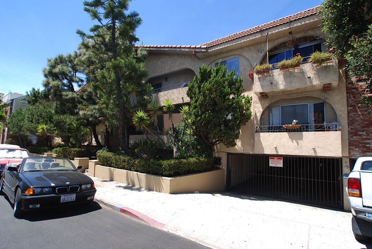Two-story stucco building with arched windows, small balconies, and a closed garage door. Black convertible parked in front.