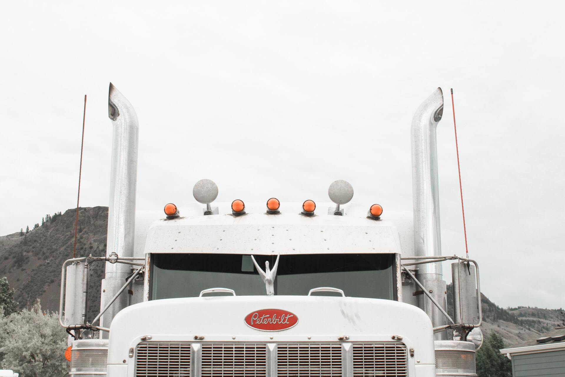 White Peterbilt semi-truck front view, with tall exhaust pipes, orange lights, and a mountain backdrop.