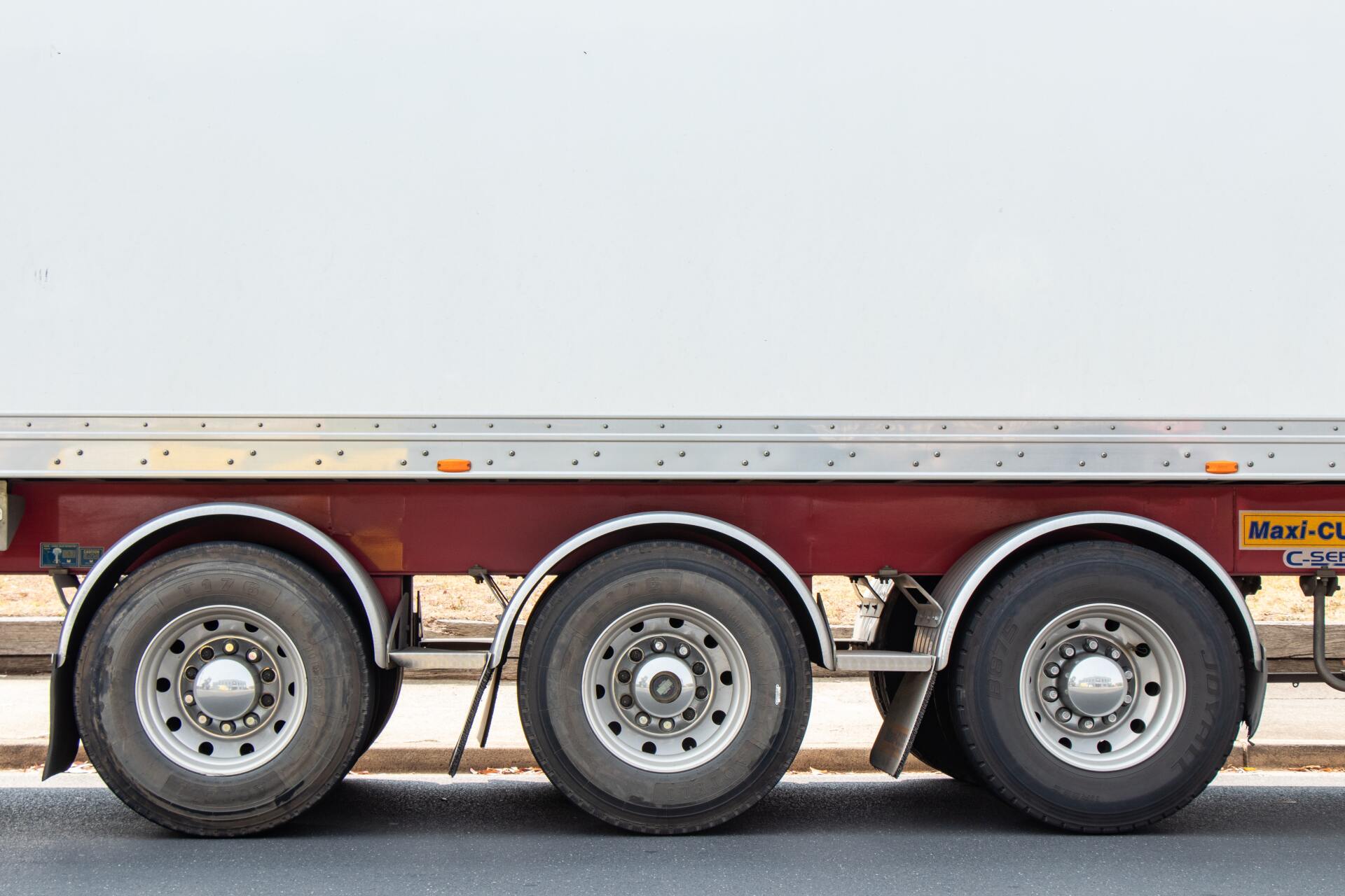 Side view of a semi-trailer truck with three rear wheels, red frame, and white cargo container.
