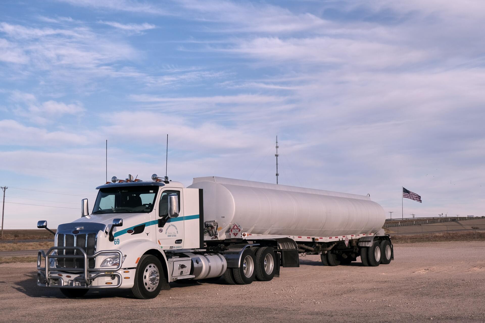 White tanker truck parked on gravel with a blue sky background. An American flag flies in the distance.