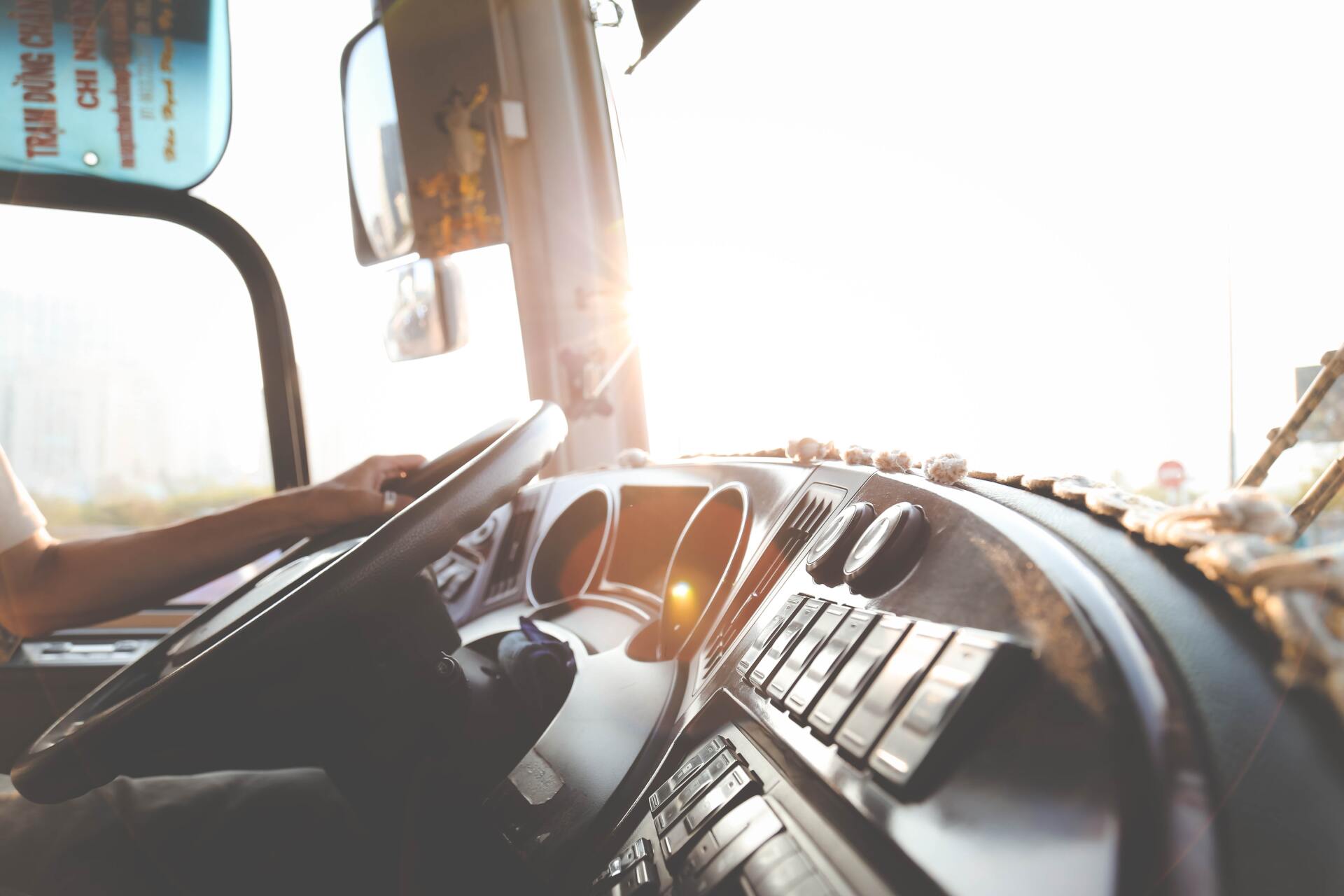 Driver's hands on steering wheel of a bus, dashboard visible. Sunlight streams in from the front.