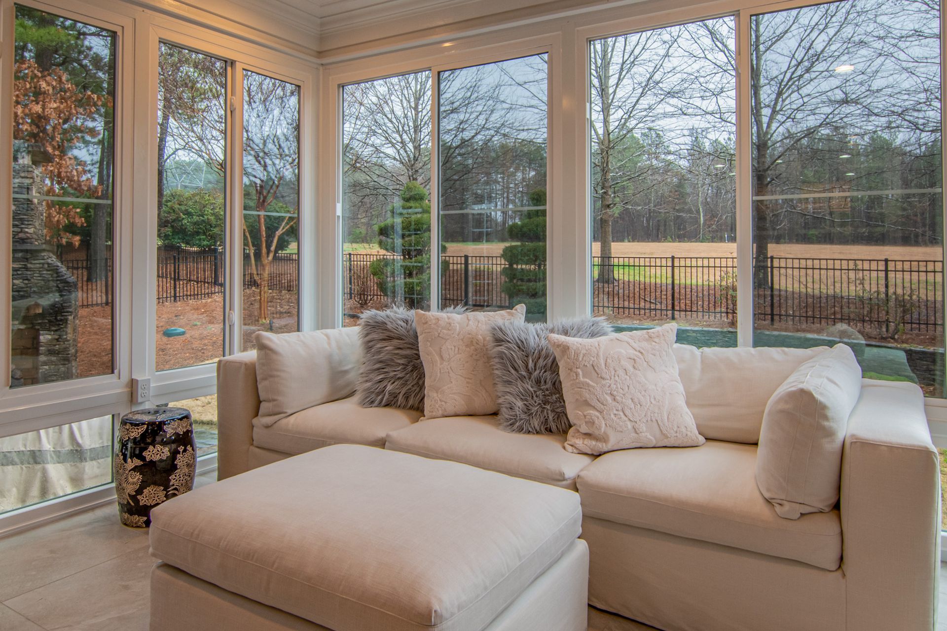 Sunroom with white sectional sofa, ottoman, and large windows overlooking a yard.