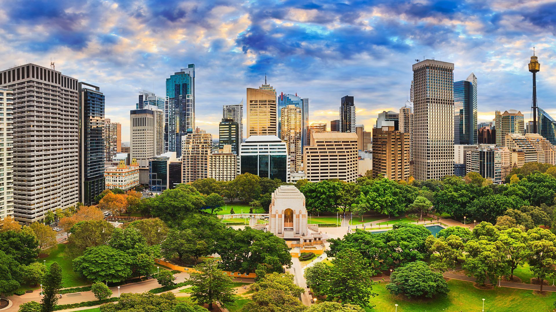 Sydney skyline with park in foreground, featuring high-rise buildings and monuments under a cloudy blue sky.
