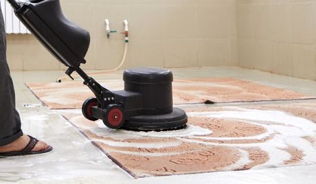 Eco Friend Carpet Clean - Person using a floor scrubber to clean a brown rug in a room with a light-colored wall.
