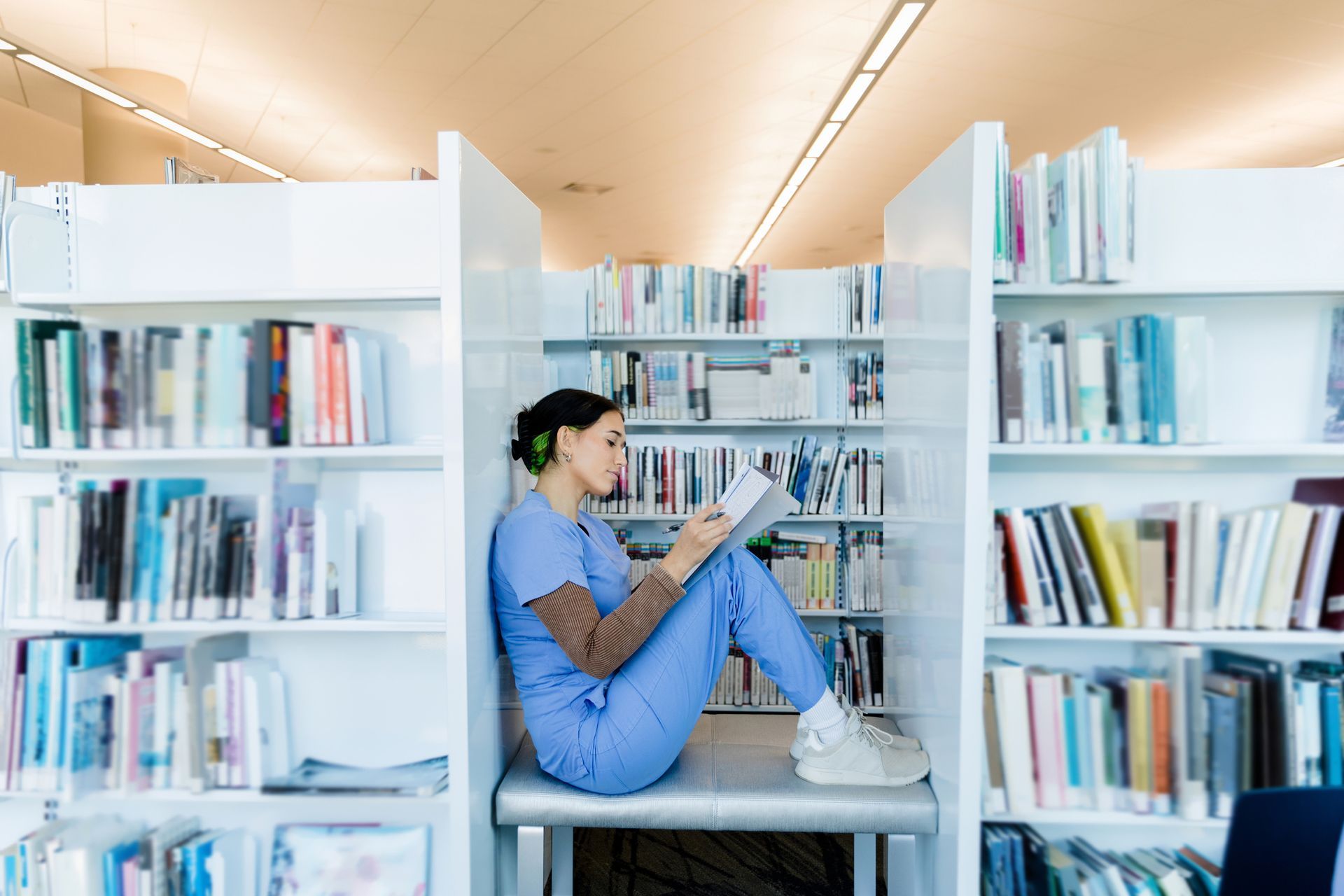 A nurse is sitting on a bench in a library reading a book.