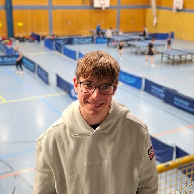 Ein junger Mann mit Brille und blauem Hemd steht vor einer gelben Wand.