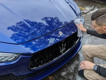A man is cleaning the front of a blue maserati with a towel.