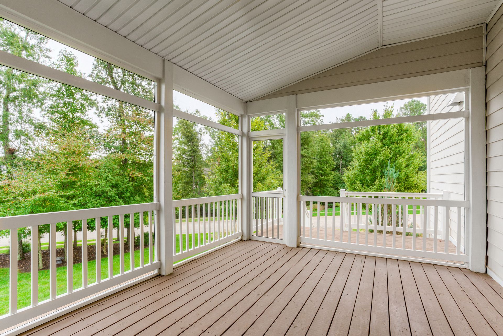 An empty screened in porch with a wooden deck and white railing.