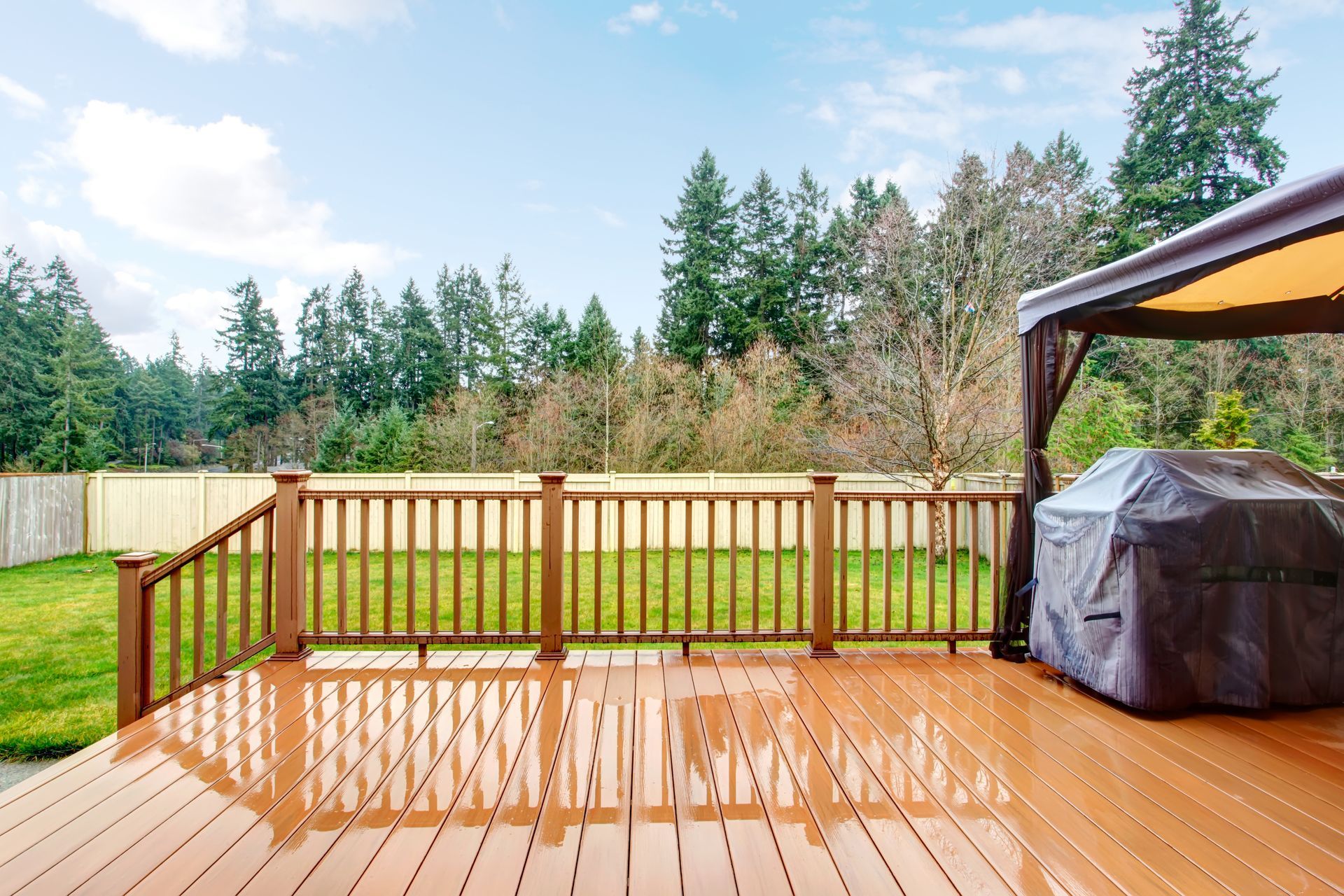 Wooden deck with railing, covered grill, and gazebo. Lawn, fence, and trees in background. Cloudy sky.