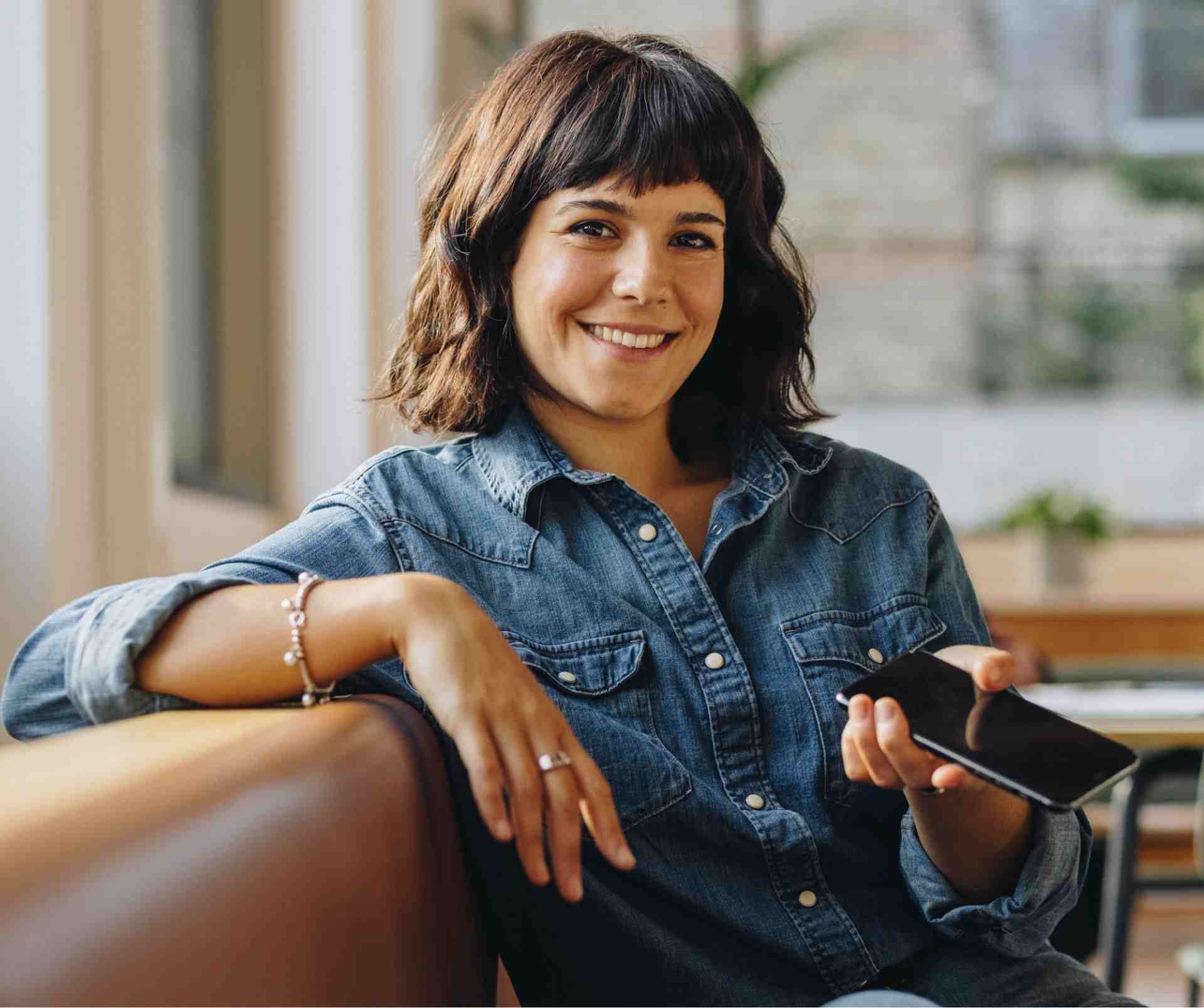 A woman in a denim shirt is sitting on a couch holding a cell phone