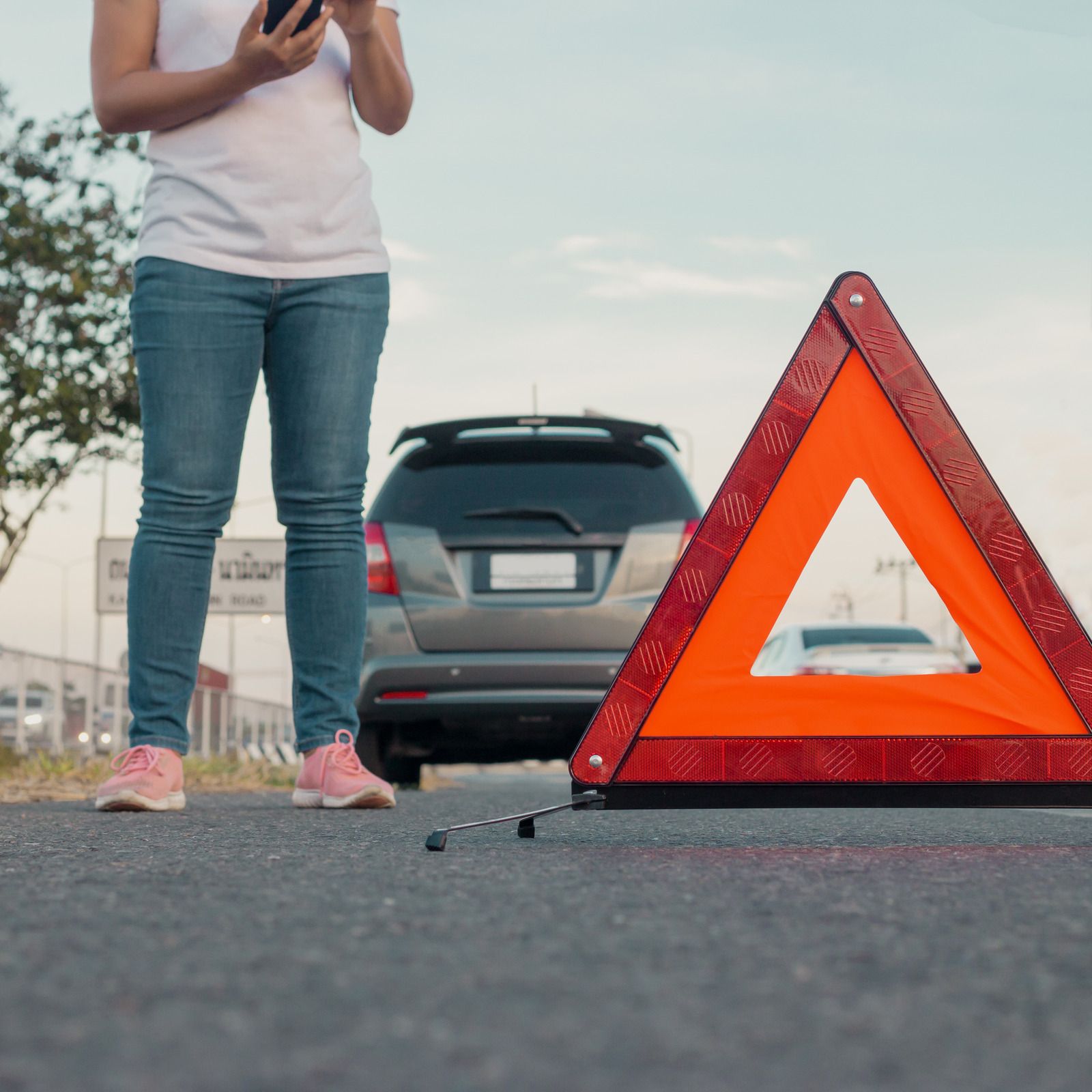 Woman standing by a car with an orange warning triangle on the road, using her phone.