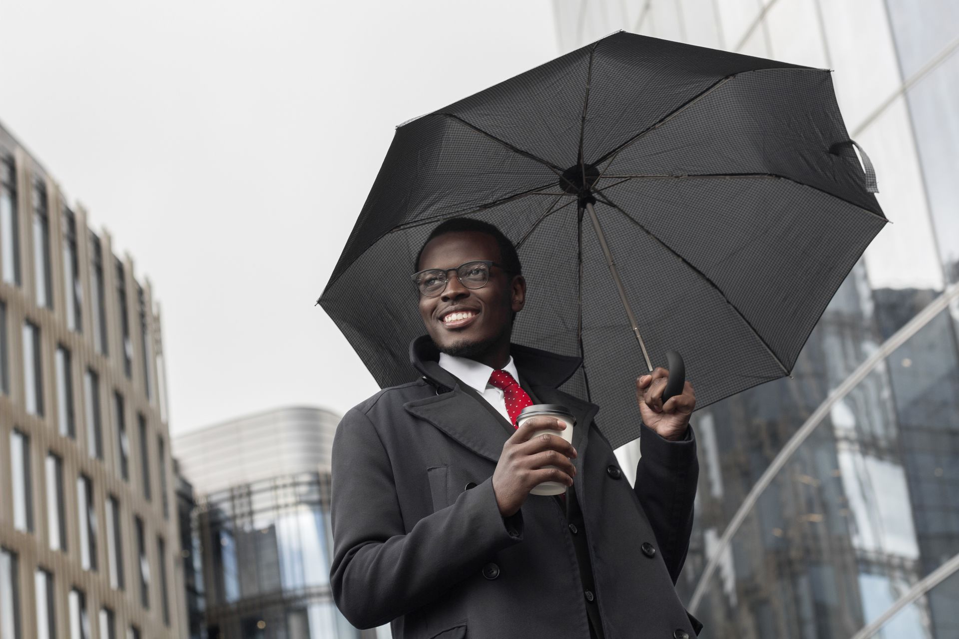 Man in suit holds umbrella and coffee cup, walking outside city buildings.