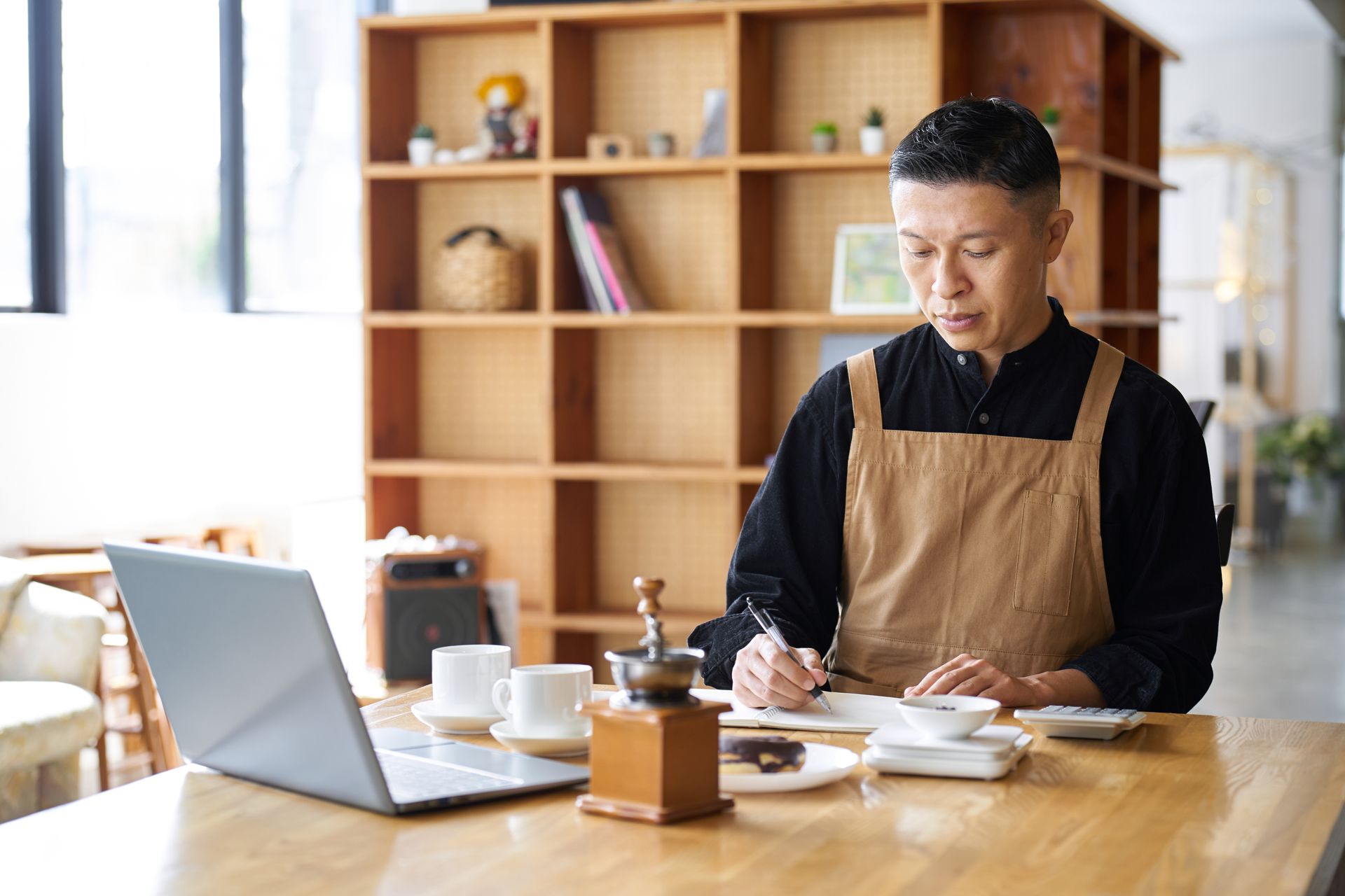 Man wearing apron working on a laptop, writing at a wooden table in a cafe.