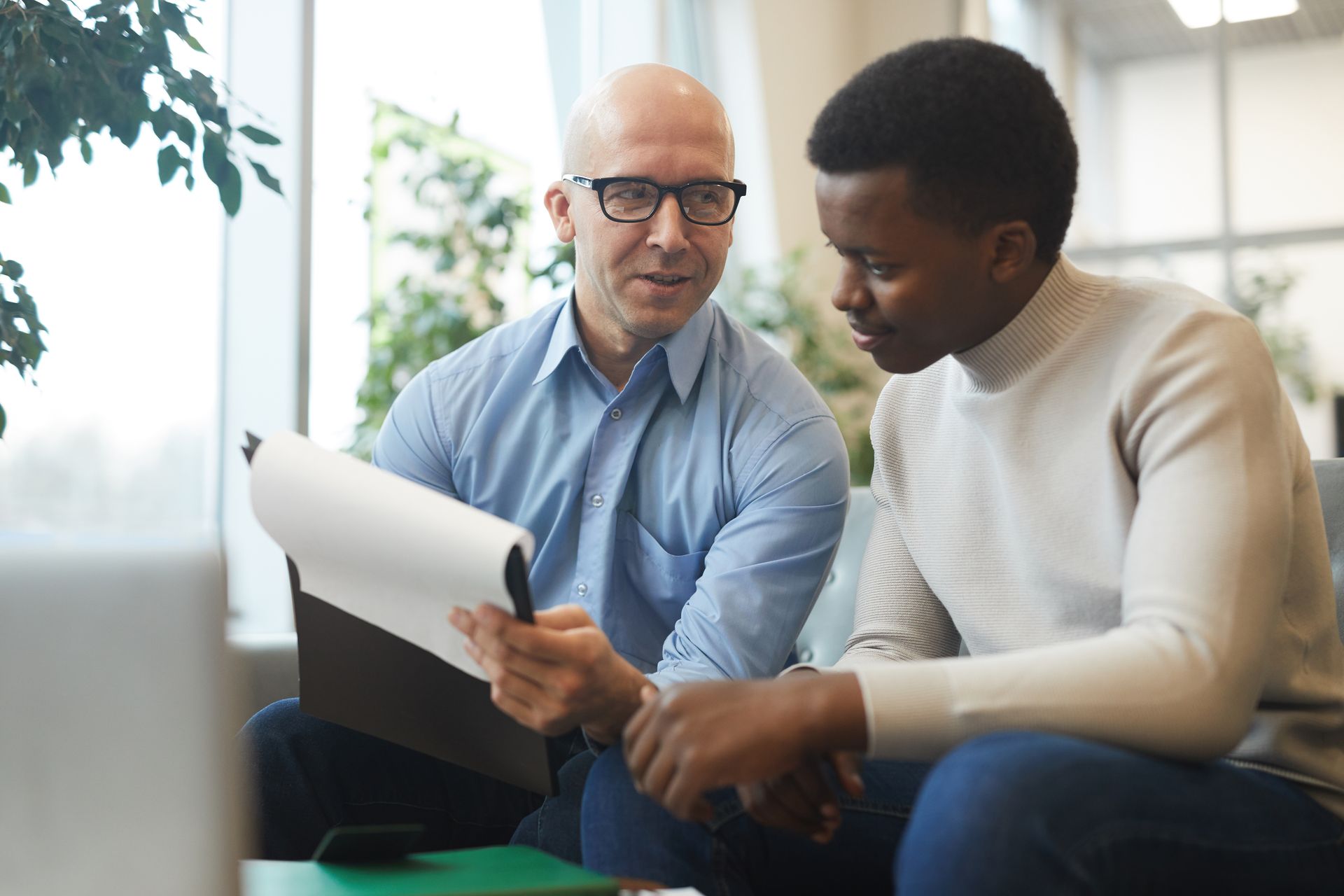 Man showing document to another man indoors; both look at the document.