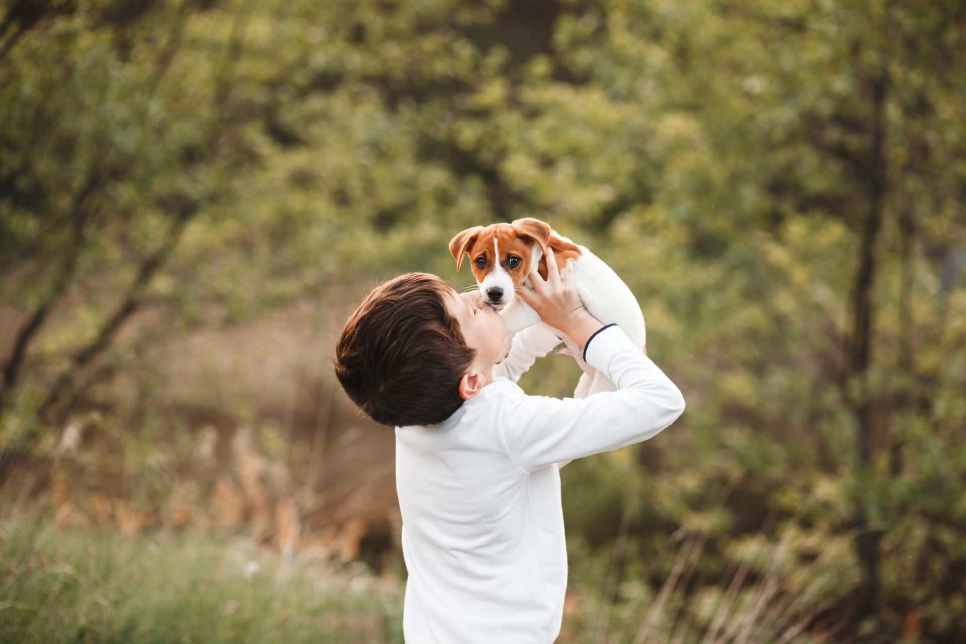 Boy holding a small dog up in the air, both looking at each other outdoors.