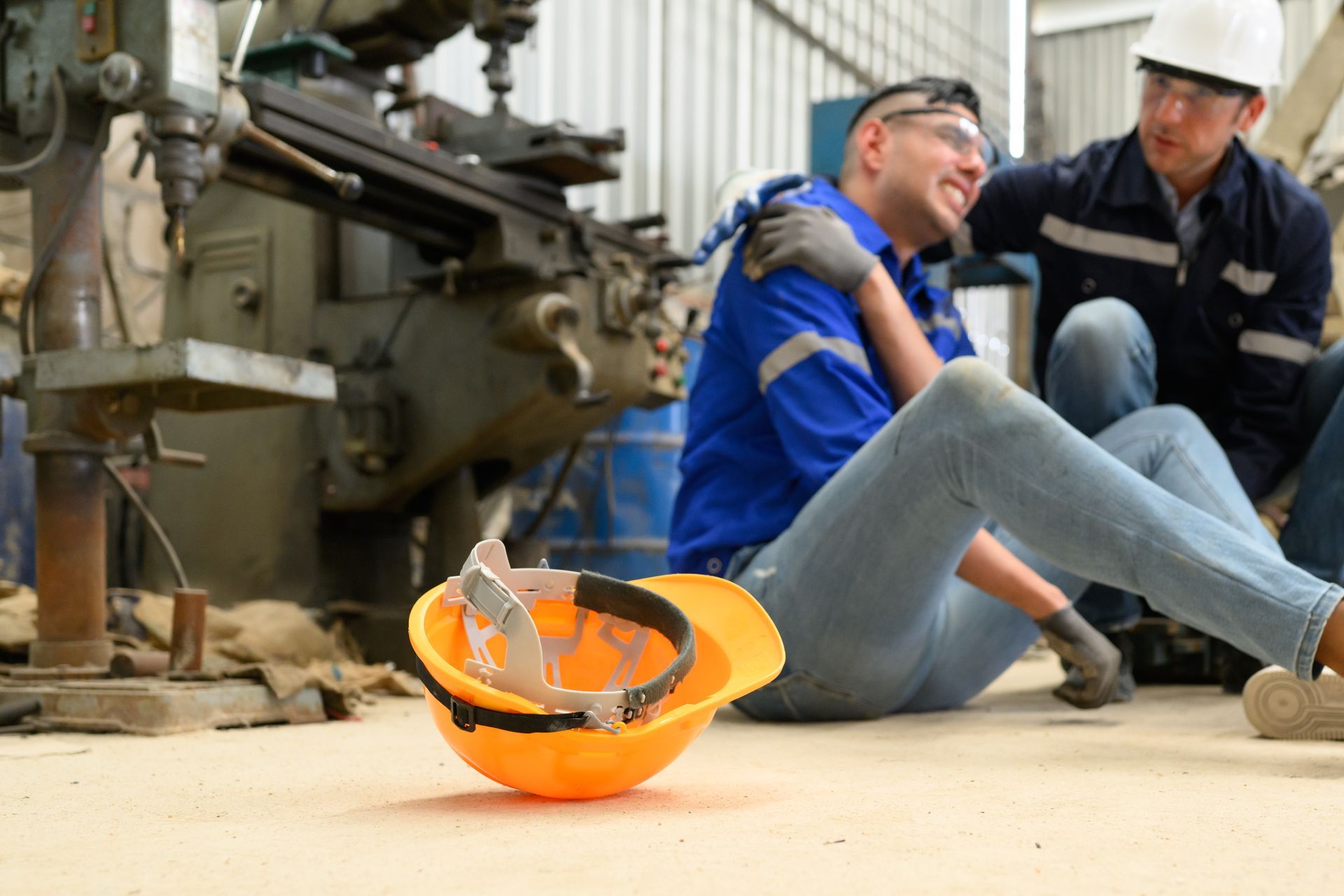 Man injured on factory floor; another worker helps. Tools, machinery in background.