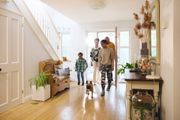 Family entering a brightly lit home, dog in front. Wooden floors, staircase, and side table with decor.