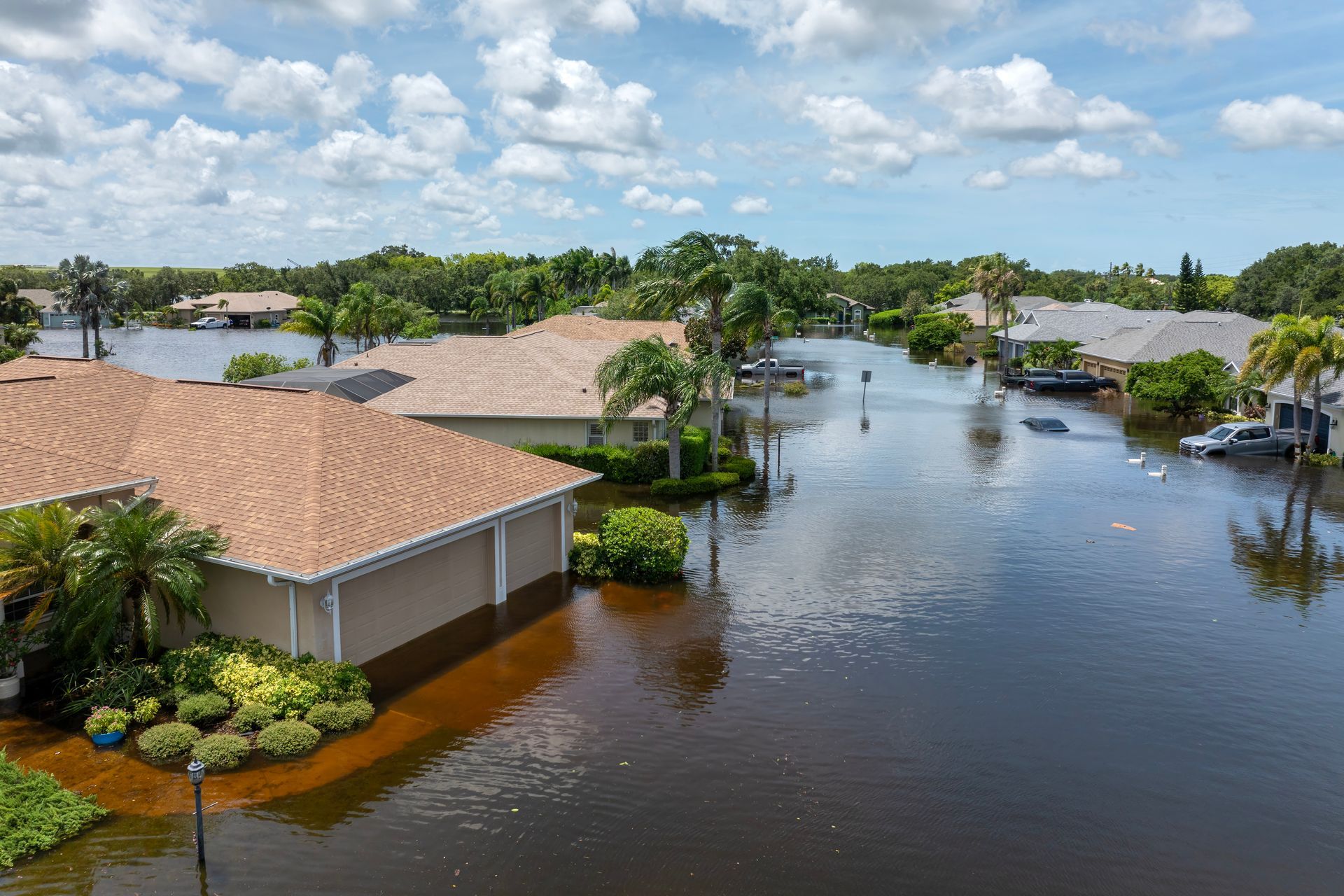 Flooded suburban neighborhood; brown floodwater surrounds houses, cars; overcast sky.