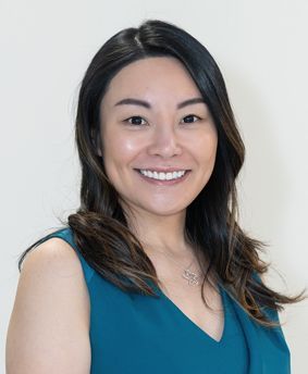 Woman with dark hair, smiling, wearing a teal top, against a white background.