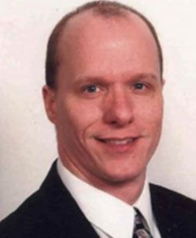 Man in dark suit smiles, head and shoulders visible against a light background.