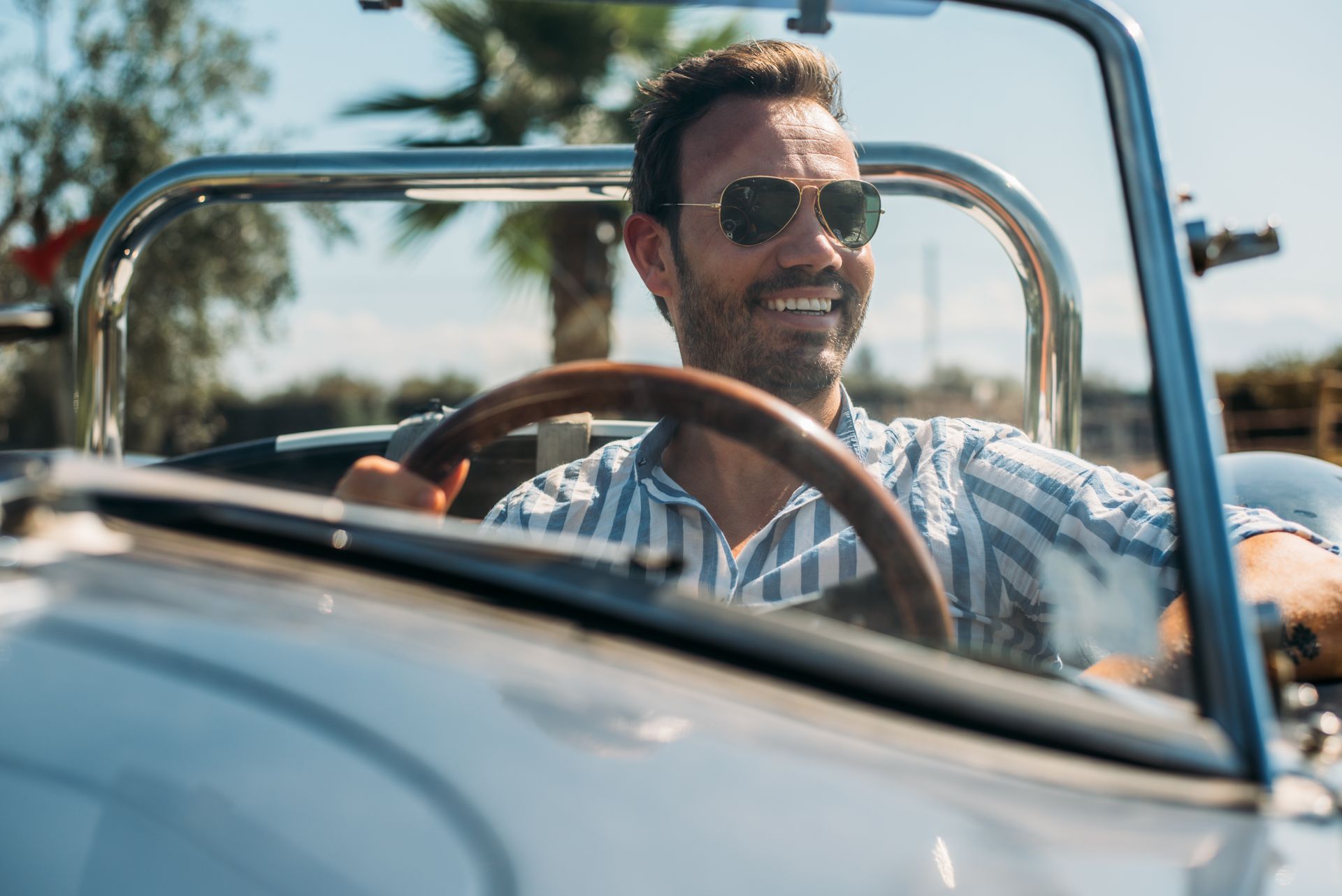 Man smiling, driving a vintage convertible car on a sunny day.