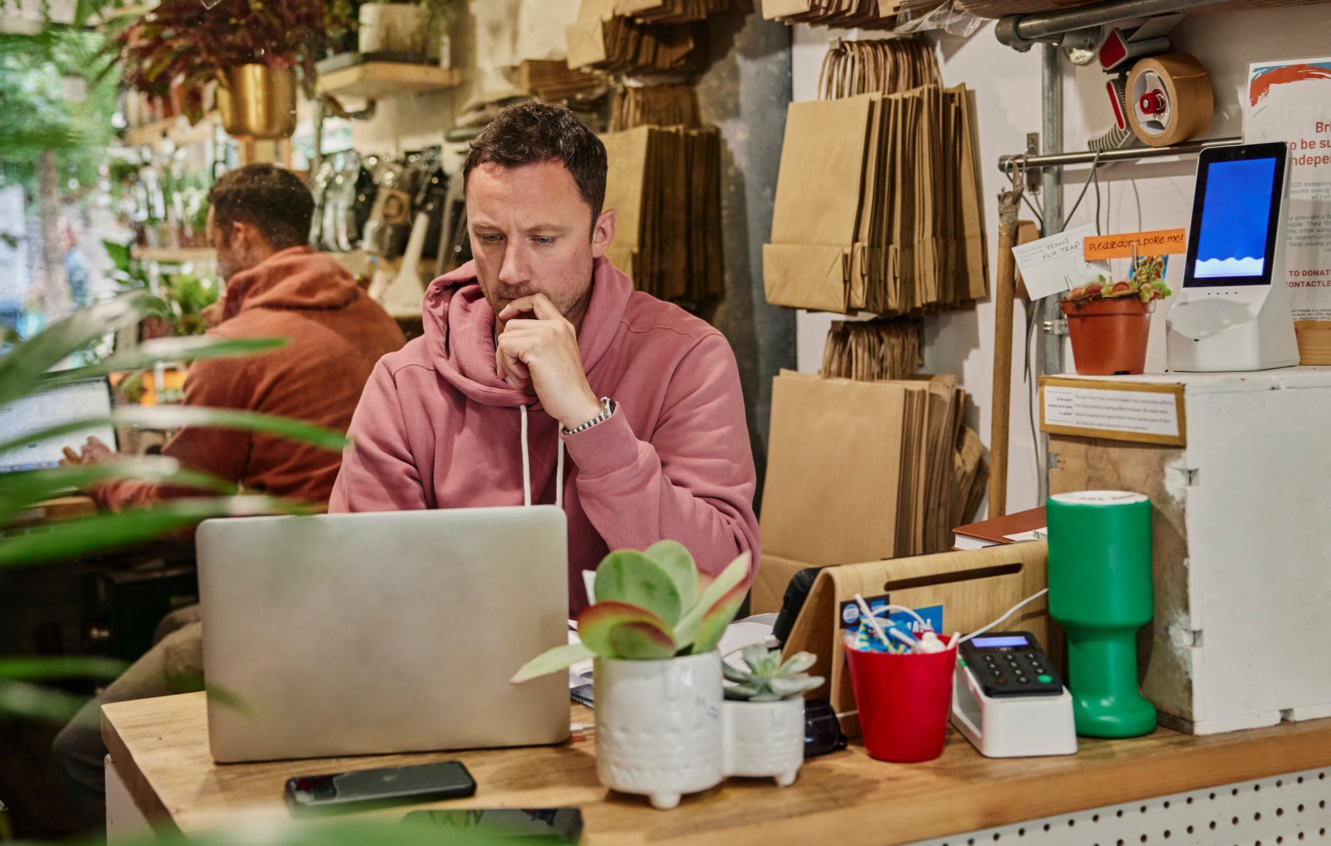 Man in pink hoodie works on a laptop at a shop counter, with another person in the background.