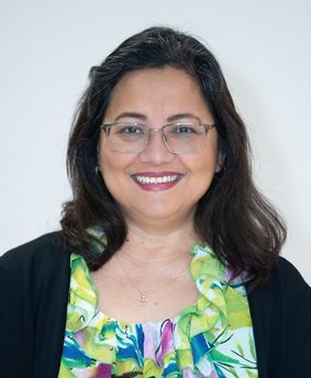 Woman with glasses, smiling, wearing a black jacket and a floral top in front of a white wall.