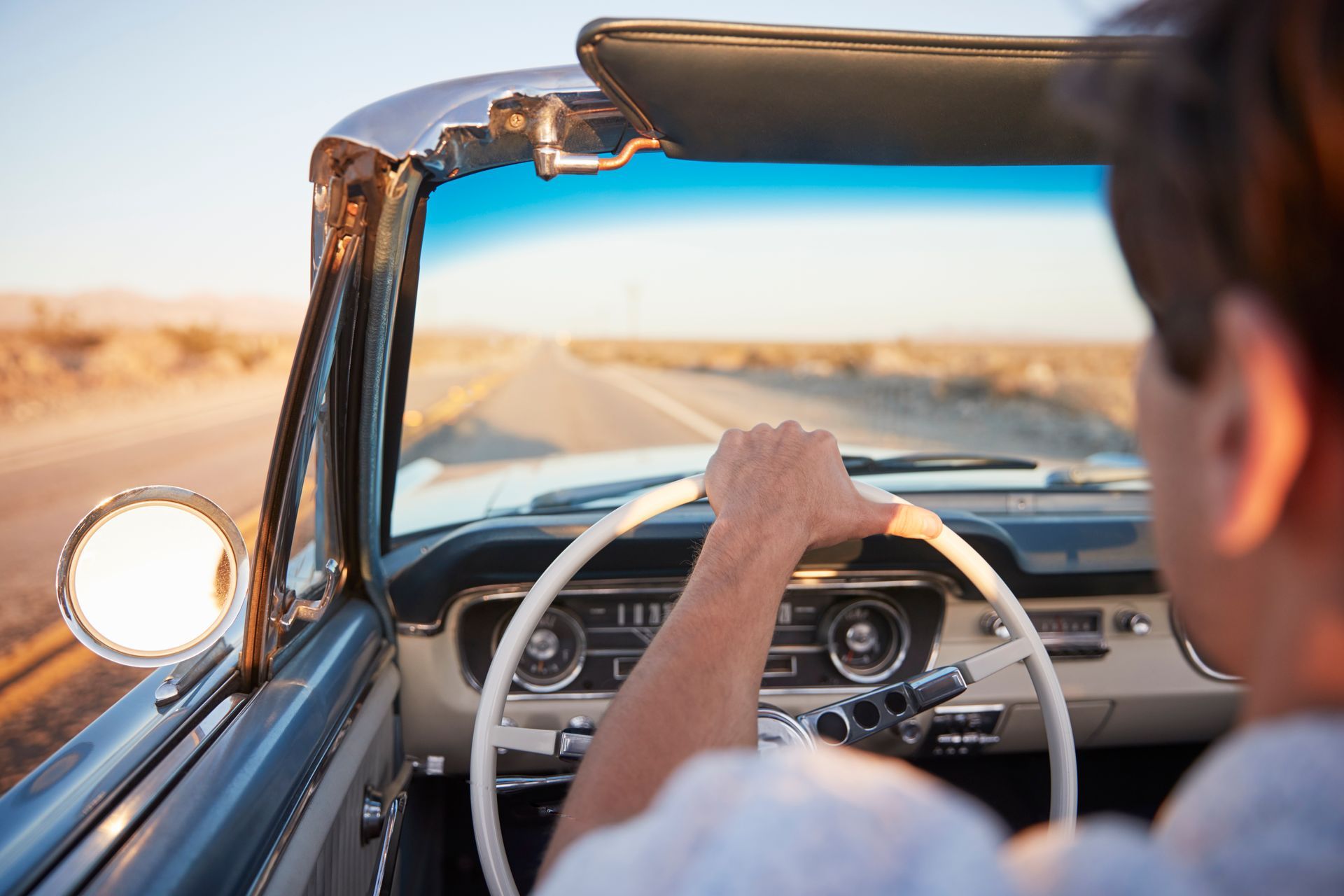 Man driving a convertible on a desert road; sunny with a blue sky.
