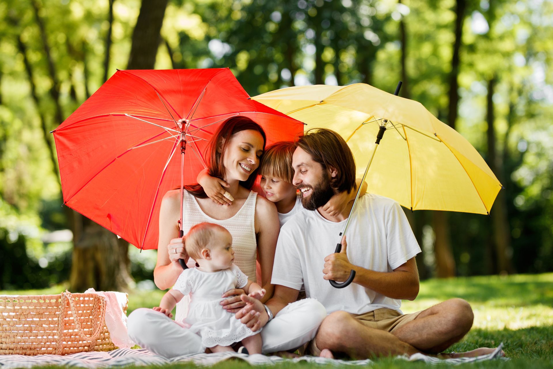 Family of four under red and yellow umbrellas, seated on a picnic blanket in a park.