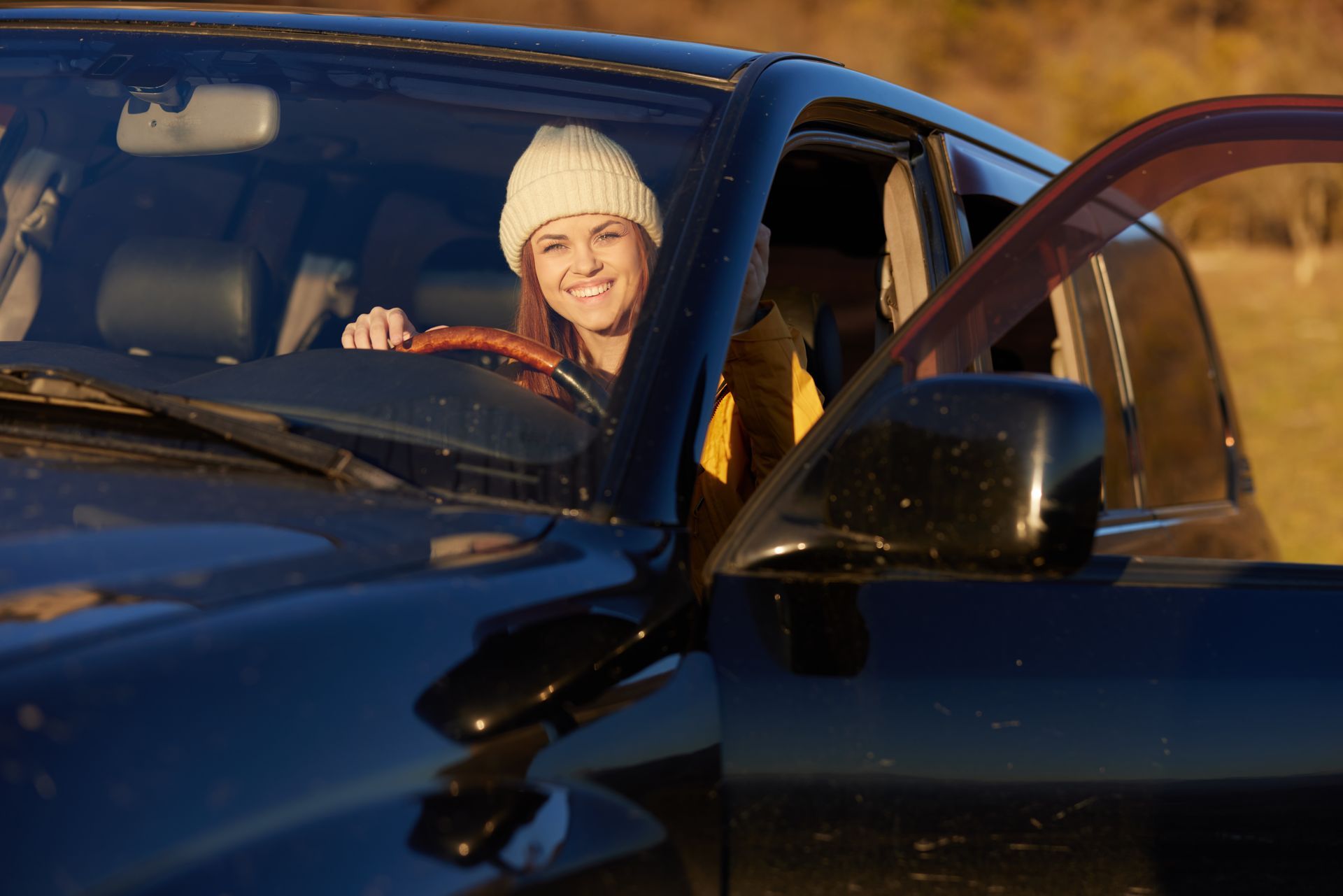 Woman in a beanie smiles from inside a black SUV, door open, setting is outdoors.