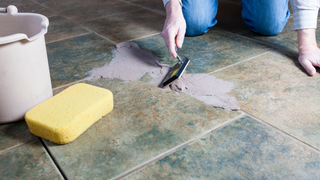 A person is cleaning a tile floor with a spatula and sponge.
