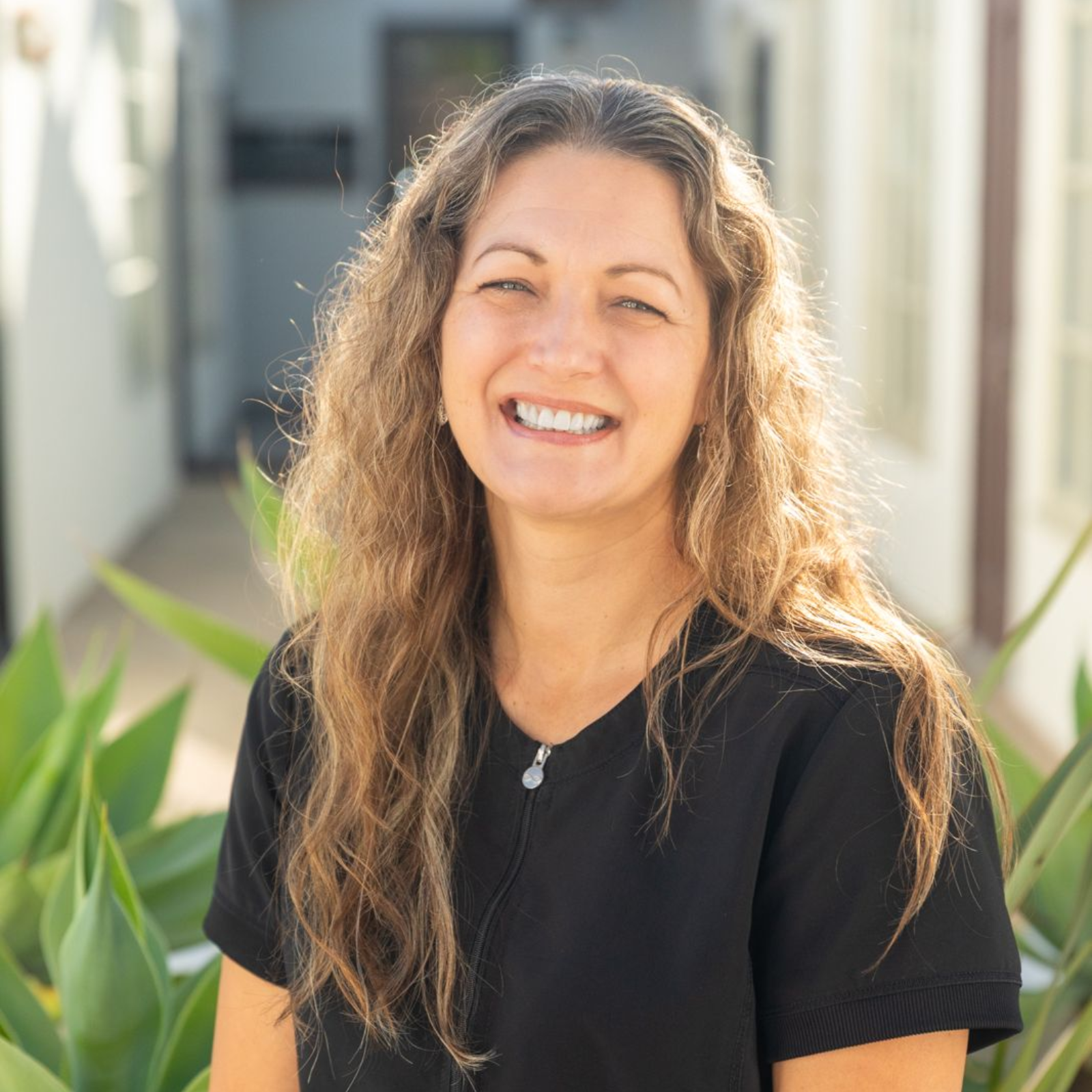 Woman with long, wavy brown hair, smiling, wearing a black top, outdoors with plants.