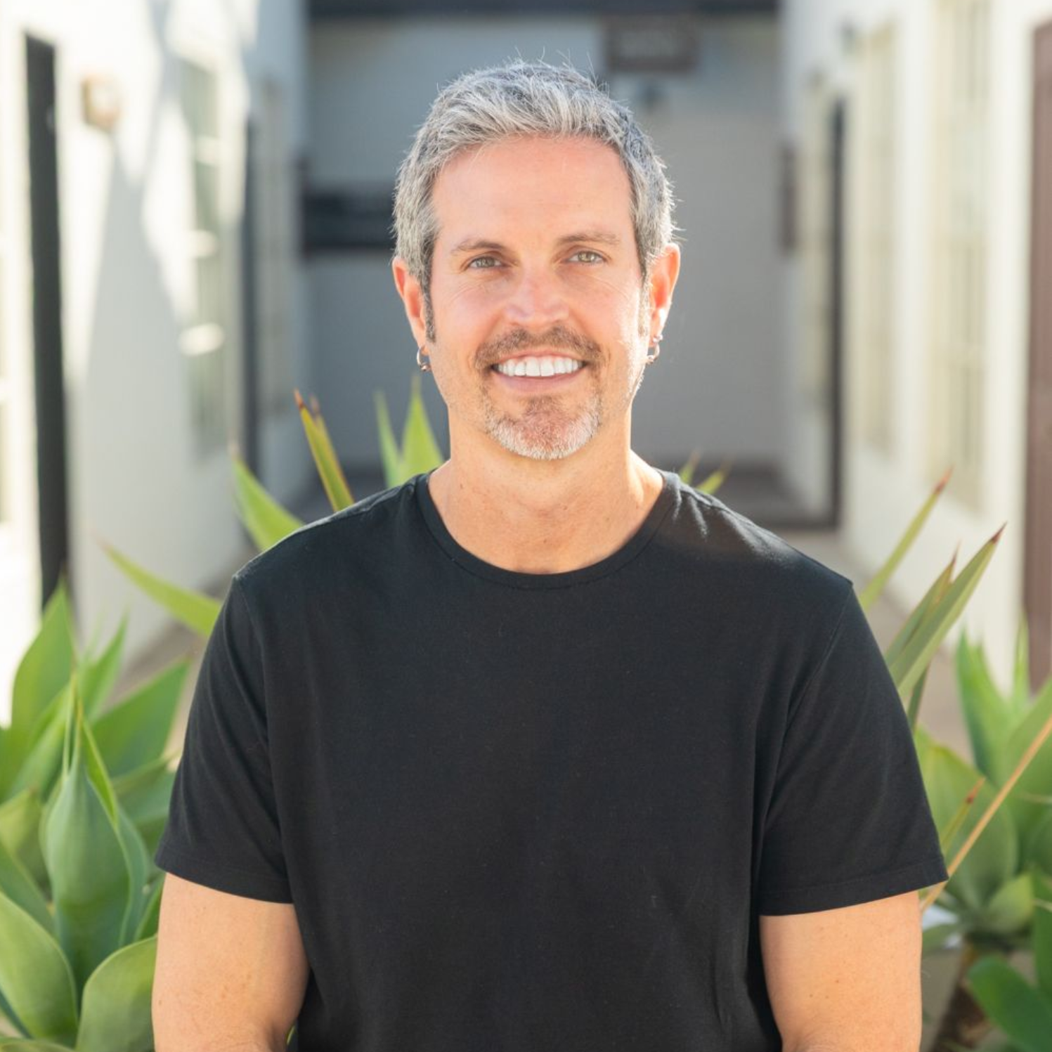 Man with gray hair smiles, wearing a black t-shirt, standing outdoors in front of a building with plants.