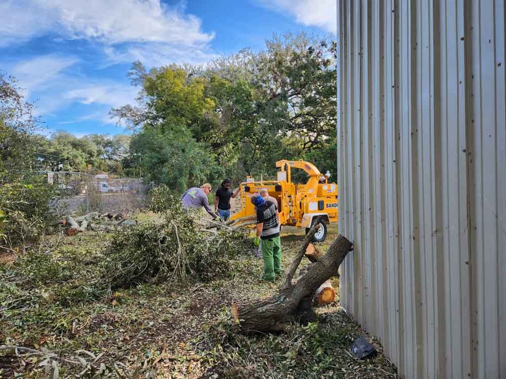 People using a wood chipper outside a building with trees in the background. Blue sky with clouds.