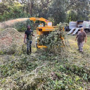 Two people feed brush into a yellow wood chipper in a grassy yard. Wood chips spray out.