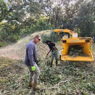 Two people feed brush into a yellow wood chipper in a wooded area.