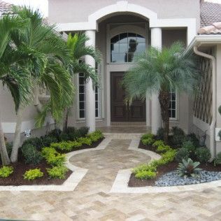 Beige house entrance with arched doorway and path, palm trees, and landscaped flower beds.
