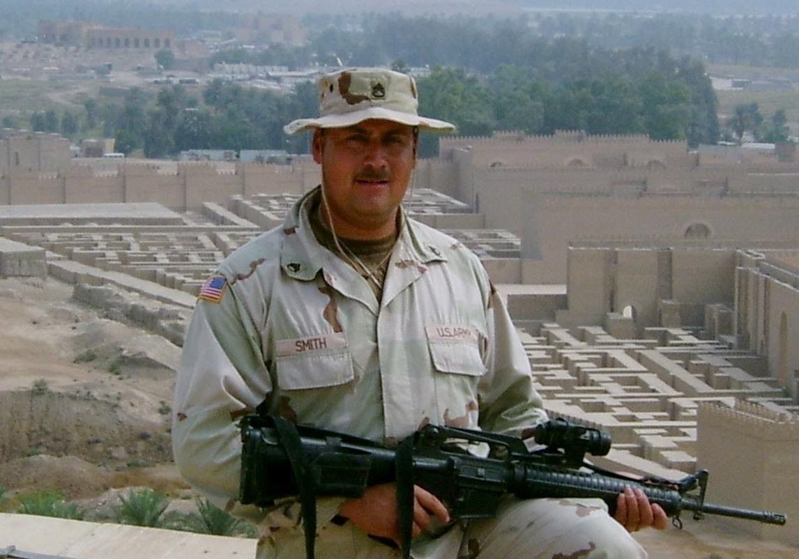 Soldier in tan uniform, holding rifle, in front of ancient ruins.