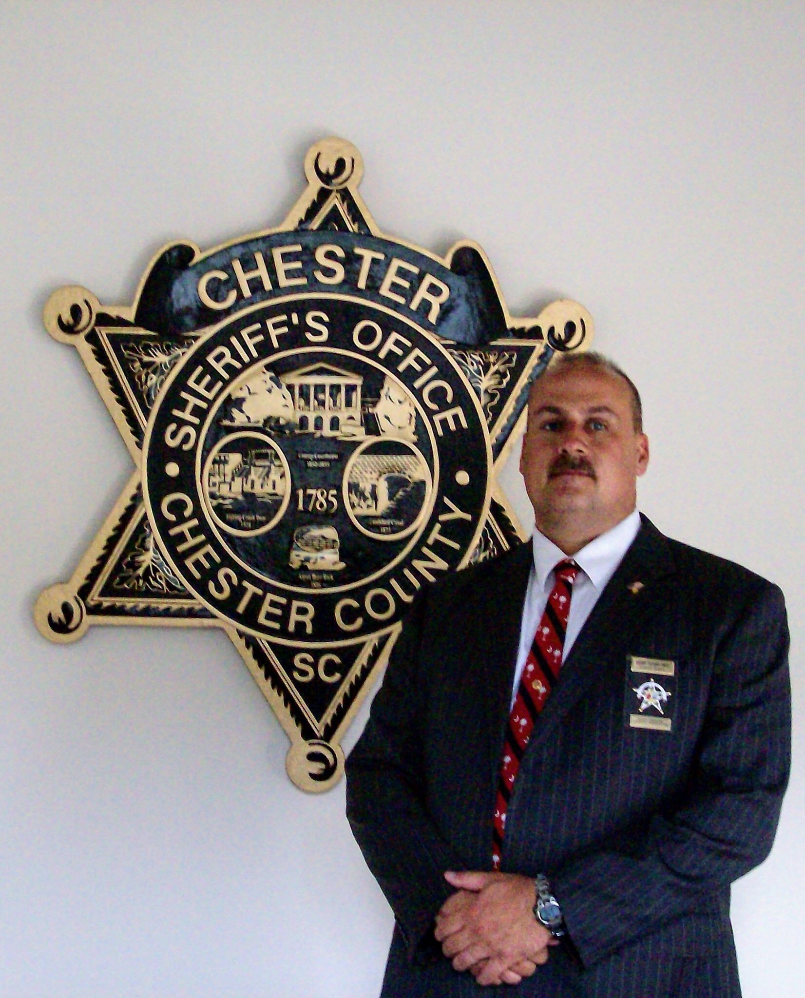 Man in suit stands in front of Chester County Sheriff's Office star badge.