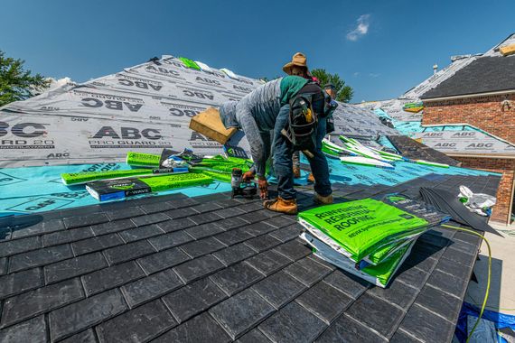 Two workers installing dark roof shingles on a house with stacked roofing materials and safety gear nearby
