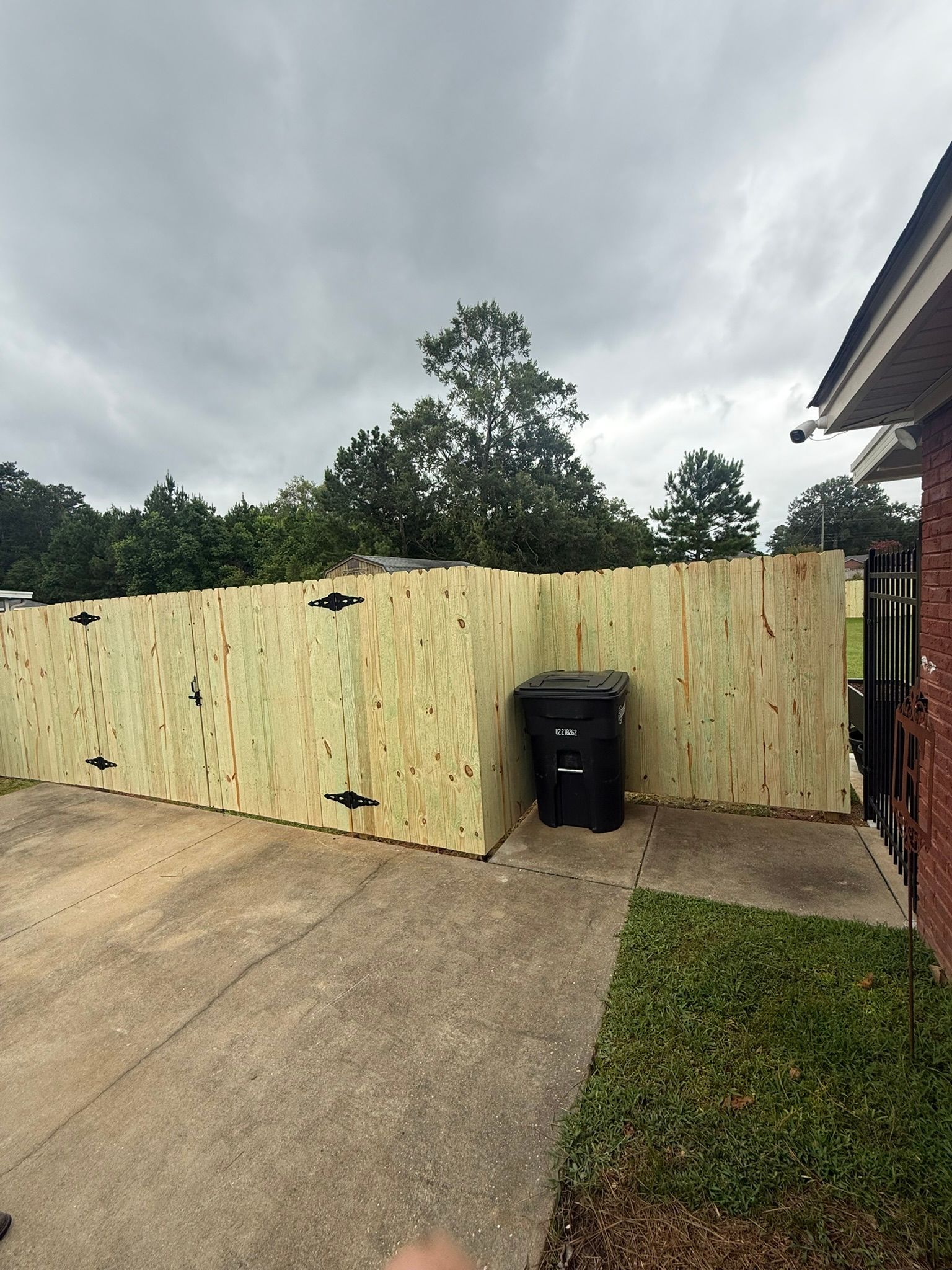 Wooden fence surrounding a black trash can on a concrete patio, next to a brick building.