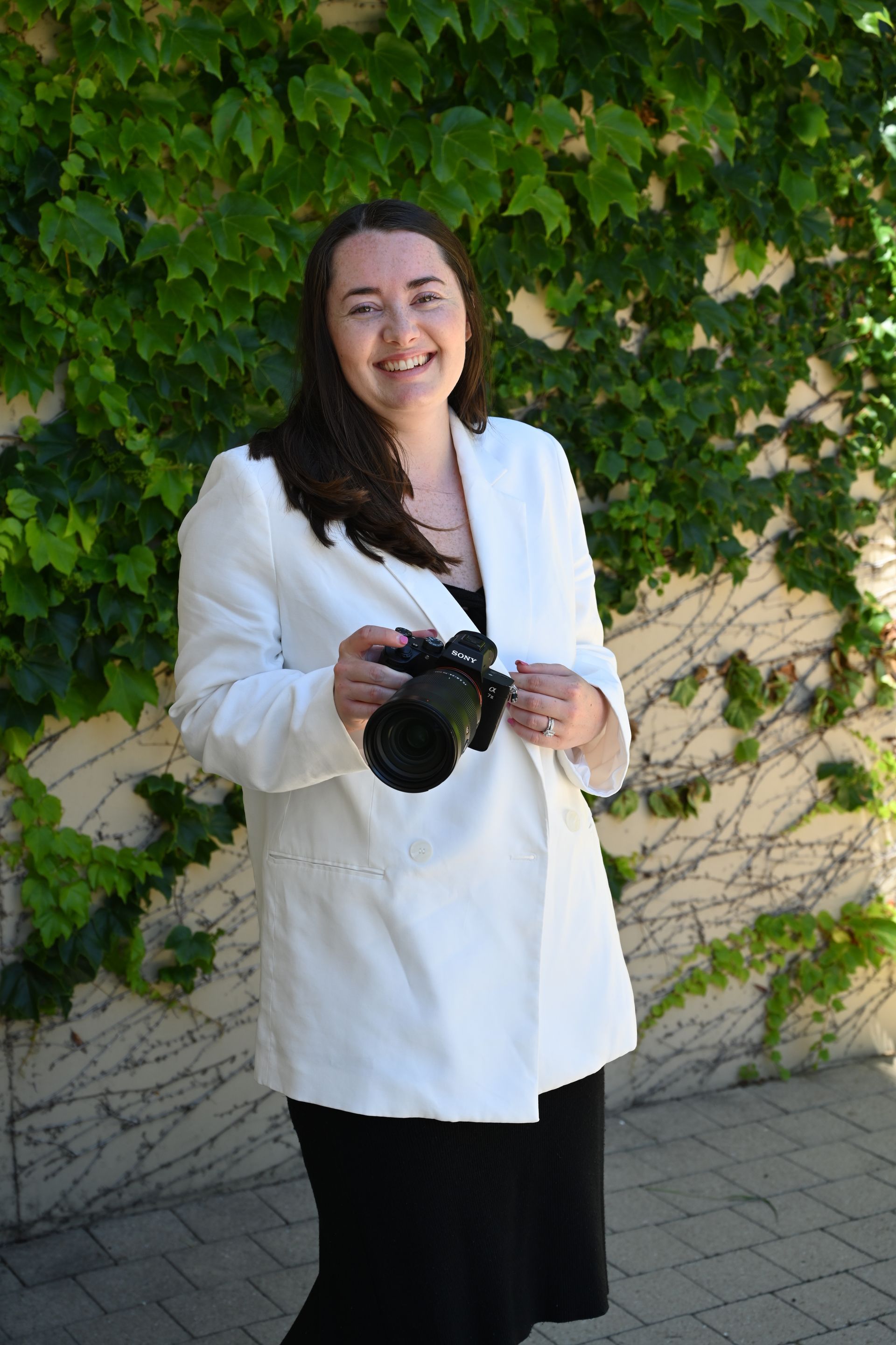 A woman in a white jacket is holding a camera in front of a ivy covered wall.