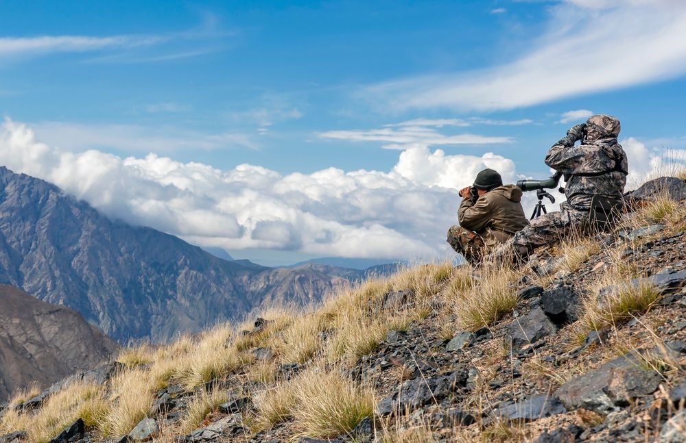 Two men are sitting on top of a mountain looking through binoculars.