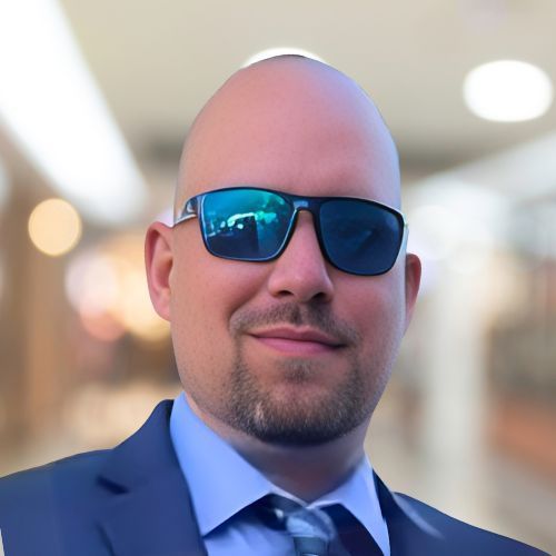 Bald man wearing sunglasses and a suit, smiling in a blurred indoor setting.